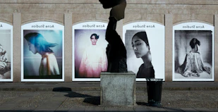 A series of large, framed photographs displayed outdoors, each featuring a different portrait. The portraits have a vintage and artistic style, with varied expressions and poses. A stone sculpture stands in the foreground, creating an interesting contrast between the three-dimensional sculpture and the flat photographic images.
