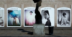A series of large, framed photographs displayed outdoors, each featuring a different portrait. The portraits have a vintage and artistic style, with varied expressions and poses. A stone sculpture stands in the foreground, creating an interesting contrast between the three-dimensional sculpture and the flat photographic images.