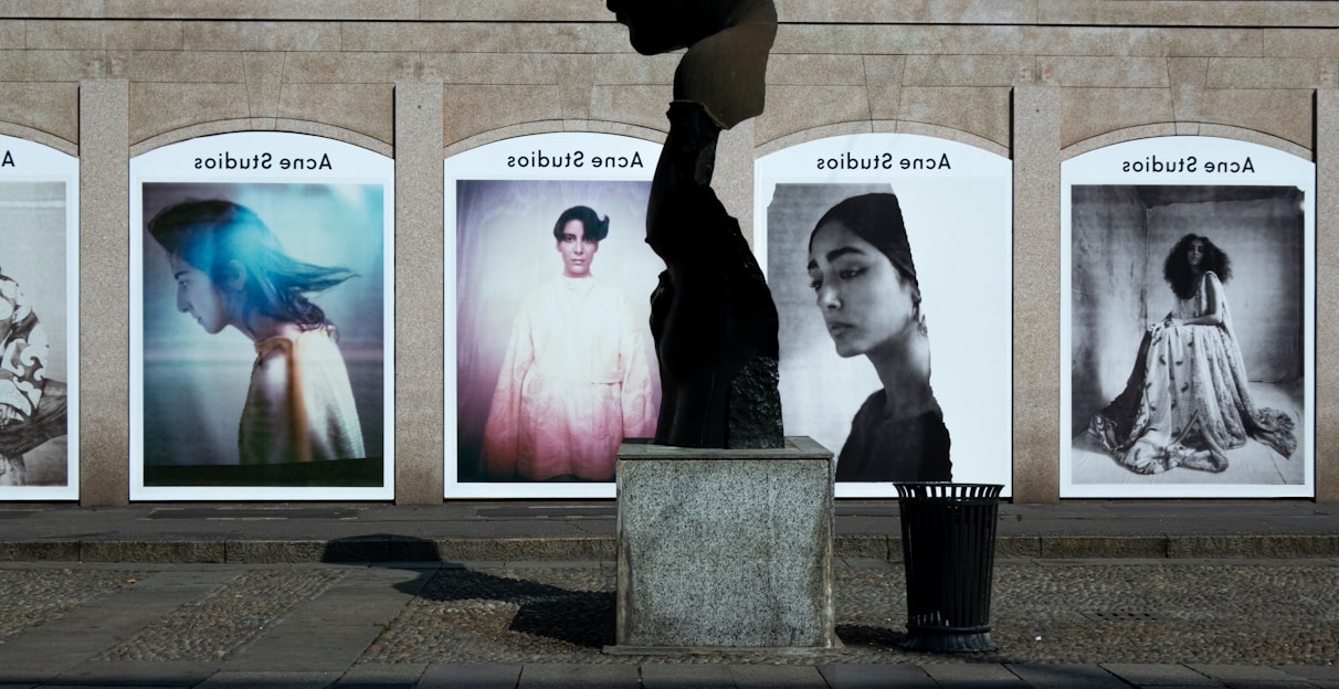A series of large, framed photographs displayed outdoors, each featuring a different portrait. The portraits have a vintage and artistic style, with varied expressions and poses. A stone sculpture stands in the foreground, creating an interesting contrast between the three-dimensional sculpture and the flat photographic images.