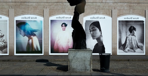 A series of large, framed photographs displayed outdoors, each featuring a different portrait. The portraits have a vintage and artistic style, with varied expressions and poses. A stone sculpture stands in the foreground, creating an interesting contrast between the three-dimensional sculpture and the flat photographic images.