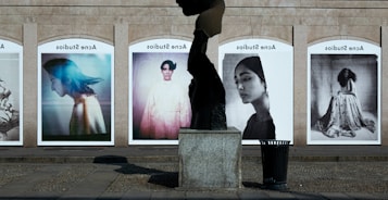 A series of large, framed photographs displayed outdoors, each featuring a different portrait. The portraits have a vintage and artistic style, with varied expressions and poses. A stone sculpture stands in the foreground, creating an interesting contrast between the three-dimensional sculpture and the flat photographic images.