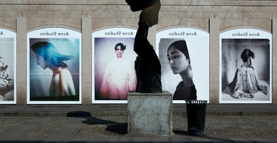 A series of large, framed photographs displayed outdoors, each featuring a different portrait. The portraits have a vintage and artistic style, with varied expressions and poses. A stone sculpture stands in the foreground, creating an interesting contrast between the three-dimensional sculpture and the flat photographic images.