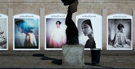 A series of large, framed photographs displayed outdoors, each featuring a different portrait. The portraits have a vintage and artistic style, with varied expressions and poses. A stone sculpture stands in the foreground, creating an interesting contrast between the three-dimensional sculpture and the flat photographic images.