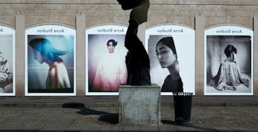 A series of large, framed photographs displayed outdoors, each featuring a different portrait. The portraits have a vintage and artistic style, with varied expressions and poses. A stone sculpture stands in the foreground, creating an interesting contrast between the three-dimensional sculpture and the flat photographic images.