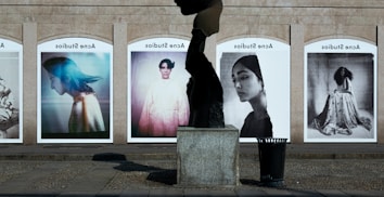 A series of large, framed photographs displayed outdoors, each featuring a different portrait. The portraits have a vintage and artistic style, with varied expressions and poses. A stone sculpture stands in the foreground, creating an interesting contrast between the three-dimensional sculpture and the flat photographic images.
