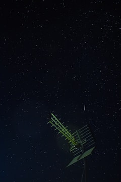 A night sky filled with numerous small stars visible against a dark background. In the foreground, a metal antenna with multiple horizontal bars is prominently positioned, slightly illuminated.