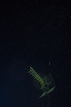 A night sky filled with numerous small stars visible against a dark background. In the foreground, a metal antenna with multiple horizontal bars is prominently positioned, slightly illuminated.
