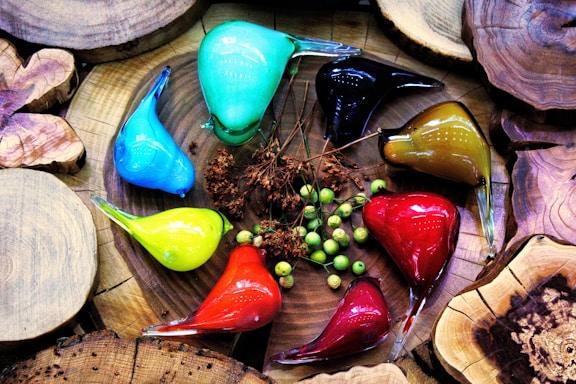 A collection of colorful glass bird sculptures arranged on a wooden surface. The birds come in various colors including cyan, blue, black, olive, yellow, red, and orange. Small green berries and dried flowers are placed among them, enhancing the natural and artistic composition. The wooden surface features cut log slices, adding a rustic element to the scene.