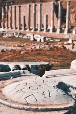 Ancient stone carvings with Greek inscriptions are in the foreground, set against a background of classical stone columns in an archaeological site.