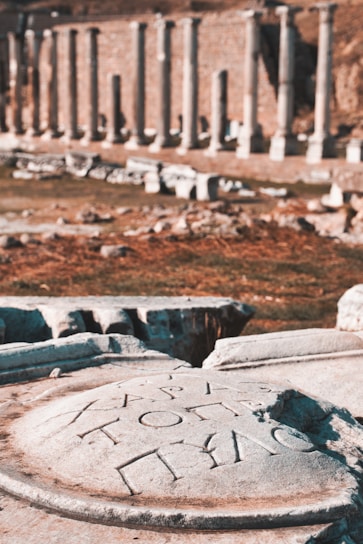 Ancient stone carvings with Greek inscriptions are in the foreground, set against a background of classical stone columns in an archaeological site.