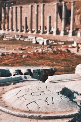 Ancient stone carvings with Greek inscriptions are in the foreground, set against a background of classical stone columns in an archaeological site.