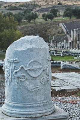 A weathered stone pillar with intricate carvings stands in the foreground of an ancient archaeological site. The surroundings feature scattered ruins, including columns and stones, with a backdrop of lush greenery and distant hills under a cloudy sky.