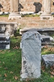 An ancient stone tablet with Greek inscriptions stands prominently in a grassy area. Surrounding it are other stone structures and remnants, likely part of a historical archaeological site. In the background, brick walls and stone columns suggest a classical architectural style.