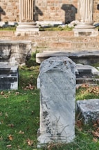 An ancient stone tablet with Greek inscriptions stands prominently in a grassy area. Surrounding it are other stone structures and remnants, likely part of a historical archaeological site. In the background, brick walls and stone columns suggest a classical architectural style.