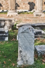 An ancient stone tablet with Greek inscriptions stands prominently in a grassy area. Surrounding it are other stone structures and remnants, likely part of a historical archaeological site. In the background, brick walls and stone columns suggest a classical architectural style.