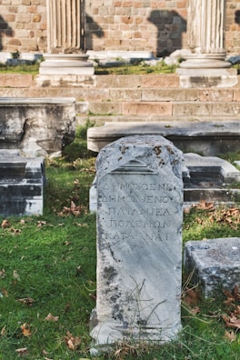 An ancient stone tablet with Greek inscriptions stands prominently in a grassy area. Surrounding it are other stone structures and remnants, likely part of a historical archaeological site. In the background, brick walls and stone columns suggest a classical architectural style.