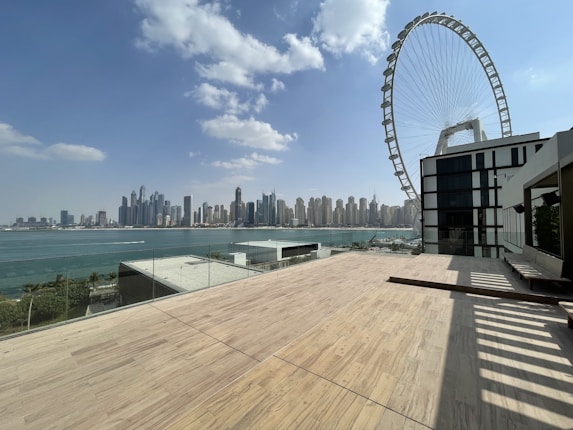 A spacious rooftop terrace overlooks a city skyline across the water, with a large Ferris wheel prominently in view. The sky is partly cloudy, and the terrace is bordered by a safety glass railing. Modern skyscrapers rise in the distance against a bright blue sky.