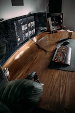 The image features a modern, minimalist workspace with a large wooden desk. Multiple computer monitors display digital content, and a mechanical keyboard with backlit keys is positioned on a mat. A gaming PC with visible internal components is on one side, and there is a potted plant adding a touch of greenery to the setup. A pair of headphones rests on the desk, and a coffee cup is visible on the side, suggesting a cozy and functional environment.