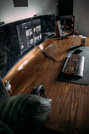 The image features a modern, minimalist workspace with a large wooden desk. Multiple computer monitors display digital content, and a mechanical keyboard with backlit keys is positioned on a mat. A gaming PC with visible internal components is on one side, and there is a potted plant adding a touch of greenery to the setup. A pair of headphones rests on the desk, and a coffee cup is visible on the side, suggesting a cozy and functional environment.