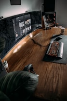 The image features a modern, minimalist workspace with a large wooden desk. Multiple computer monitors display digital content, and a mechanical keyboard with backlit keys is positioned on a mat. A gaming PC with visible internal components is on one side, and there is a potted plant adding a touch of greenery to the setup. A pair of headphones rests on the desk, and a coffee cup is visible on the side, suggesting a cozy and functional environment.