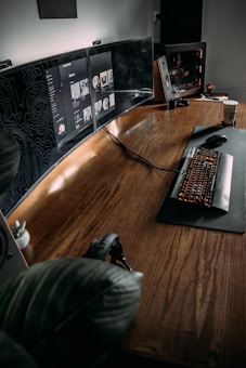 The image features a modern, minimalist workspace with a large wooden desk. Multiple computer monitors display digital content, and a mechanical keyboard with backlit keys is positioned on a mat. A gaming PC with visible internal components is on one side, and there is a potted plant adding a touch of greenery to the setup. A pair of headphones rests on the desk, and a coffee cup is visible on the side, suggesting a cozy and functional environment.