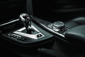 The image features the interior of a car, focusing on the gear shift and a control knob. The gear shift has a sleek design with metallic and black accents, indicative of a luxury or high-performance vehicle. Beside it is an intricate control system, including a dial with surrounding buttons, all set against a carbon fiber surface for a modern and sophisticated look.