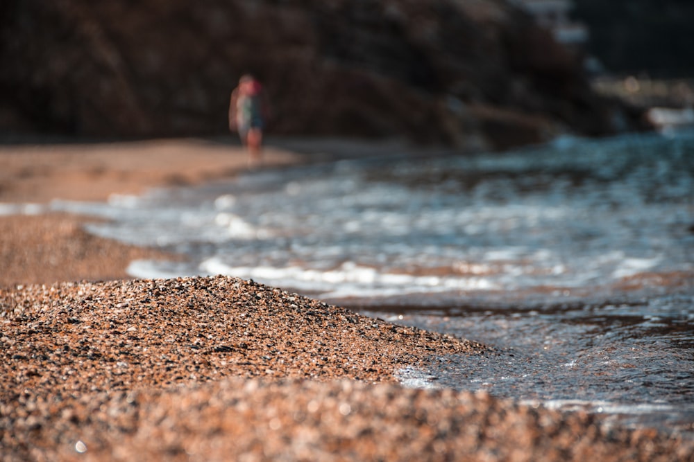 personne en chemise rouge et jean bleu debout sur le sable brun près du plan d’eau