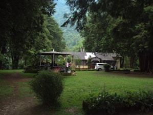 A quaint house is nestled among tall, lush trees in a serene natural setting. In front of the house, a well-trimmed lawn and a parked white car are visible. Nearby, a covered gazebo with people sitting underneath adds a cozy touch to the scene. The surrounding greenery and distant hills create a peaceful and secluded atmosphere.