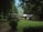 A quaint house is nestled among tall, lush trees in a serene natural setting. In front of the house, a well-trimmed lawn and a parked white car are visible. Nearby, a covered gazebo with people sitting underneath adds a cozy touch to the scene. The surrounding greenery and distant hills create a peaceful and secluded atmosphere.