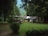 A quaint house is nestled among tall, lush trees in a serene natural setting. In front of the house, a well-trimmed lawn and a parked white car are visible. Nearby, a covered gazebo with people sitting underneath adds a cozy touch to the scene. The surrounding greenery and distant hills create a peaceful and secluded atmosphere.