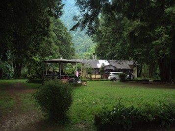 A quaint house is nestled among tall, lush trees in a serene natural setting. In front of the house, a well-trimmed lawn and a parked white car are visible. Nearby, a covered gazebo with people sitting underneath adds a cozy touch to the scene. The surrounding greenery and distant hills create a peaceful and secluded atmosphere.