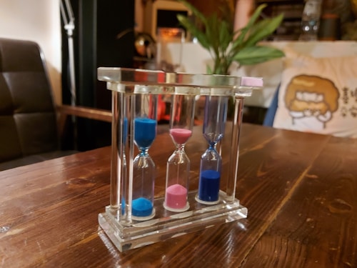 A wooden table holds a transparent three-hourglass set containing blue and pink sand. In the background, indoor plants and a cushion featuring a cartoon design add a cozy atmosphere.