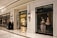 An upscale store interior with elegant white walls and a glossy marble floor. The left side features a storefront with a window displaying a mannequin in a luxurious purple and gold gown. Inside the store, a display of handbags is visible with warm lighting. Four people are standing outside, dressed in dark attire, with one pushing a stroller.