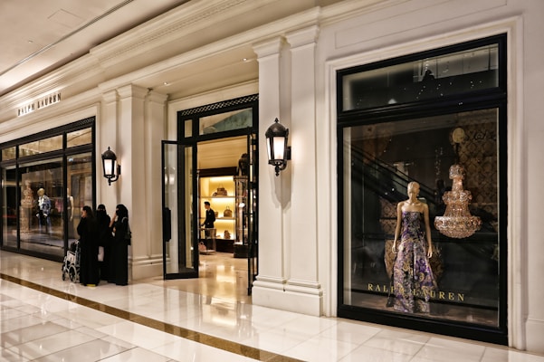 An upscale store interior with elegant white walls and a glossy marble floor. The left side features a storefront with a window displaying a mannequin in a luxurious purple and gold gown. Inside the store, a display of handbags is visible with warm lighting. Four people are standing outside, dressed in dark attire, with one pushing a stroller.