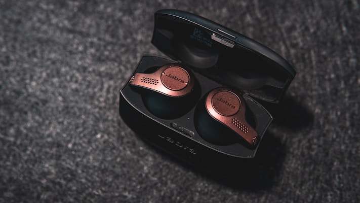 A pair of wireless earbuds with an elegant, metallic finish is placed inside an open charging case. The surface beneath has a textured, dark appearance, enhancing the sleek look of the earbuds.