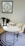 A modern workspace featuring a white oval desk with a gold frame, accompanied by a cream upholstered chair. On the desk are a pair of decorative black vases with green plants, a stylish lamp, and an open magazine. The floor is covered by a circular rug with concentric black and white rings. A framed abstract artwork with a circular pattern is hung on the wall above the desk.
