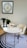 A modern workspace featuring a white oval desk with a gold frame, accompanied by a cream upholstered chair. On the desk are a pair of decorative black vases with green plants, a stylish lamp, and an open magazine. The floor is covered by a circular rug with concentric black and white rings. A framed abstract artwork with a circular pattern is hung on the wall above the desk.