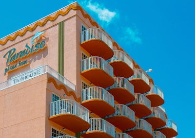 A multi-story hotel building with a distinctive wave-like architectural design. The structure has numerous balconies with white railings, contrasting with the building&rsquo;s peach-colored exterior. The sky in the background is clear and bright blue. The signage on the building reads 'Paradise Plaza Inn.'