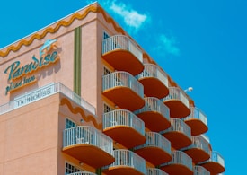 A multi-story hotel building with a distinctive wave-like architectural design. The structure has numerous balconies with white railings, contrasting with the building’s peach-colored exterior. The sky in the background is clear and bright blue. The signage on the building reads 'Paradise Plaza Inn.'