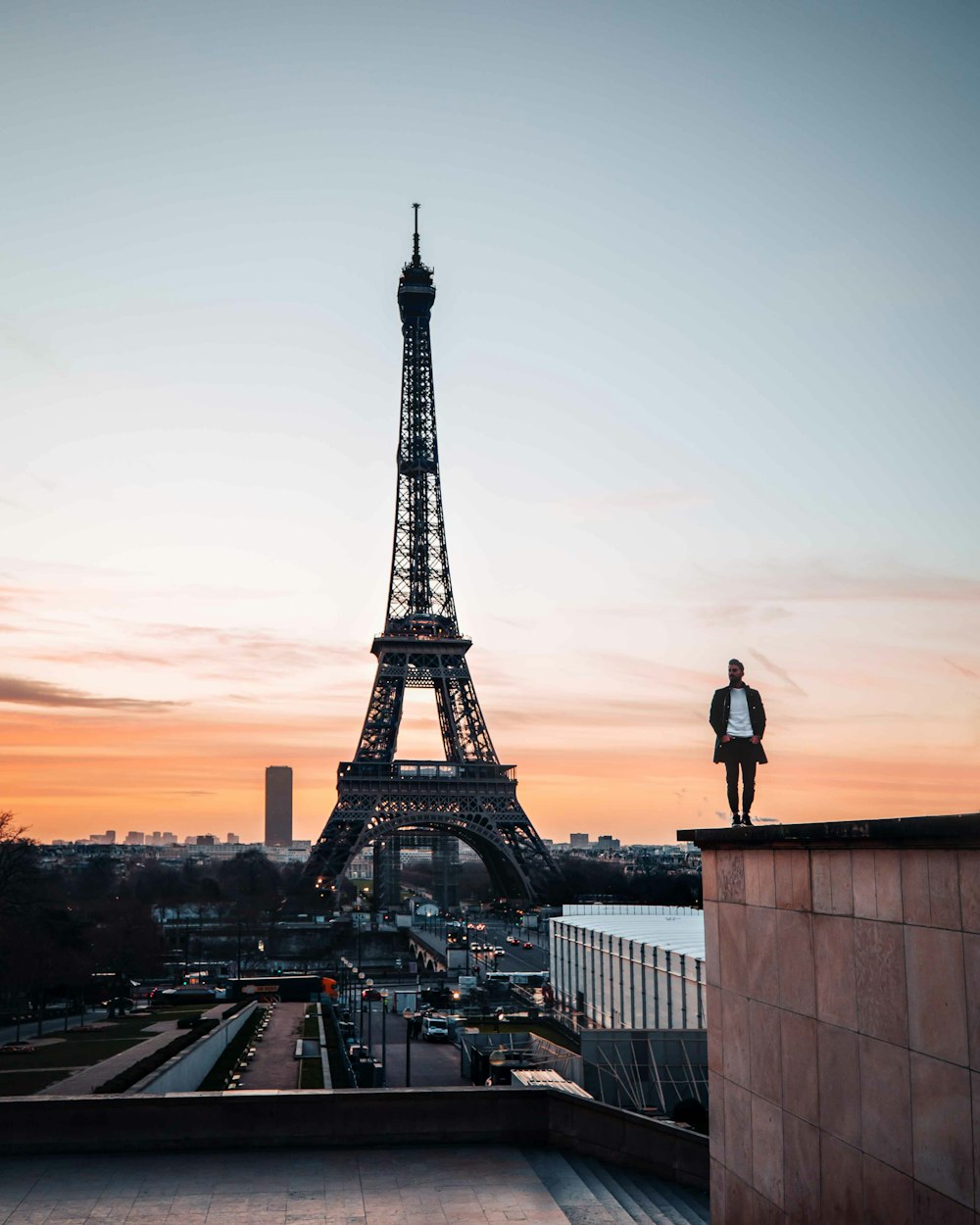 man in black jacket standing on top of building looking at eiffel tower during sunset