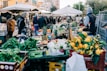 A bustling outdoor marketplace with a variety of fruits and vegetables displayed on tables. Shoppers dressed in winter clothing are browsing and purchasing goods. Stalls are covered with white canopies and surrounded by crates filled with fresh produce like lettuce, oranges, and pineapples.