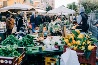 A bustling outdoor marketplace with a variety of fruits and vegetables displayed on tables. Shoppers dressed in winter clothing are browsing and purchasing goods. Stalls are covered with white canopies and surrounded by crates filled with fresh produce like lettuce, oranges, and pineapples.