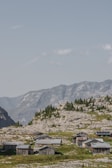 A cluster of rustic wooden cabins is scattered across a rocky hillside, surrounded by patches of green grass and sparse trees. In the background, impressive mountain peaks rise under a clear, blue sky with a few fluffy clouds.