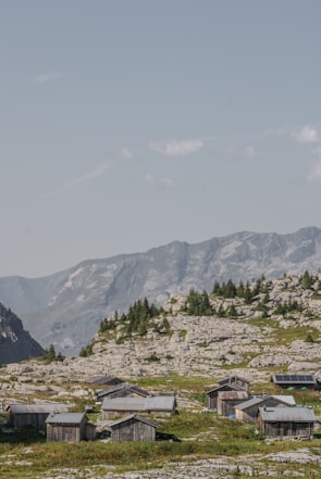 A cluster of rustic wooden cabins is scattered across a rocky hillside, surrounded by patches of green grass and sparse trees. In the background, impressive mountain peaks rise under a clear, blue sky with a few fluffy clouds.