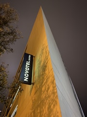 A tall, triangular building facade with a modern design is illuminated by warm, ambient lighting. The NOVOTEL Suites logo is prominently displayed on a black sign. The evening sky is overcast, and tree branches with sparse leaves create a natural silhouette against the building.
