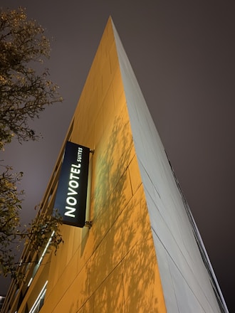 A tall, triangular building facade with a modern design is illuminated by warm, ambient lighting. The NOVOTEL Suites logo is prominently displayed on a black sign. The evening sky is overcast, and tree branches with sparse leaves create a natural silhouette against the building.