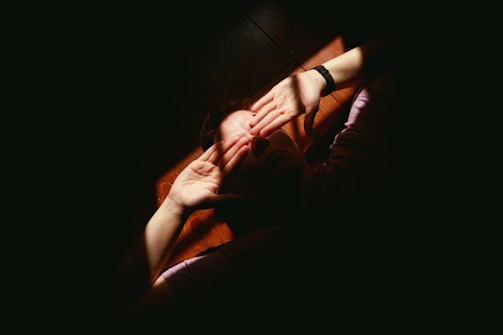 A person lying down with hands covering their face, bathed in a warm stream of light cutting through darkness. The focus is on the hands and facial expression partially hidden by shadows.