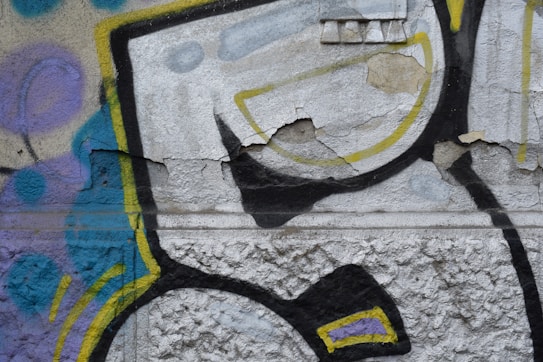 A textured wall features vibrant graffiti artwork with a mix of geometric and abstract patterns. The paint is slightly cracked and peeling, revealing layers beneath. Colors such as yellow, blue, black, and purple create dynamic shapes and lines.