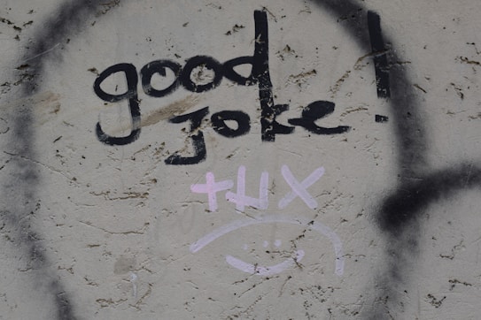 Graffiti on a textured wall displays the words 'good joke!' in bold black paint. Below it, the word 'thx' and a smiley face are drawn in light pink.