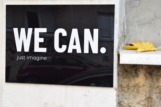 A black signboard with large white text that reads 'WE CAN.' and smaller text below that says 'just imagine'. To the right of the sign, a single yellow leaf rests on a stone ledge. In the background, there is a faint reflection of a red car.